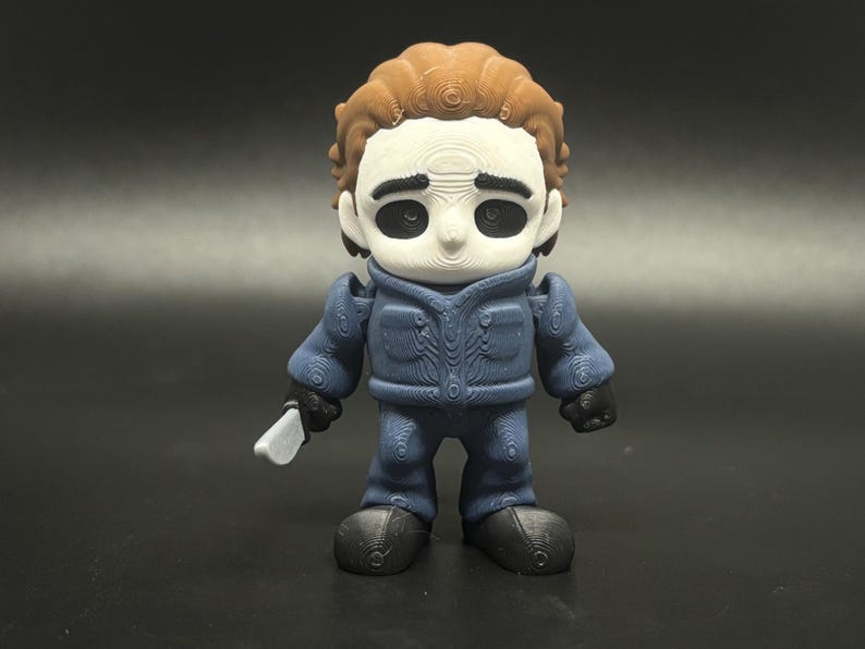 May include: A 3D-printed figure of a horror movie character. The figure has a white face, brown hair, and is wearing a blue jumpsuit and black gloves and shoes. It is holding a silver knife.