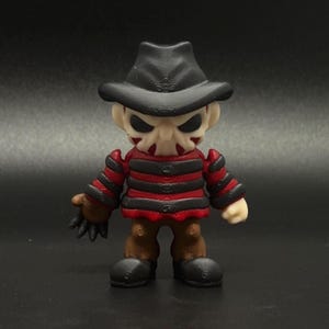 Pode incluir: Uma pequena figura com listras pretas e vermelhas, um chap&eacute;u preto e uma m&aacute;scara branca. A figura tem uma express&atilde;o amea&ccedil;adora e segura uma faca na m&atilde;o direita.