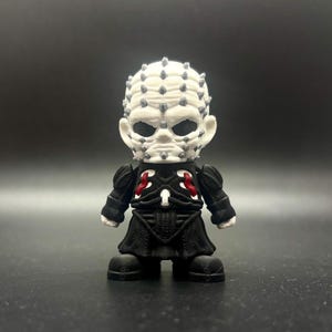 May include: A small, stylized figurine of a character with a white head covered in gray pins. The figure wears a black outfit with red accents and black boots. The figurine is a collectible item.