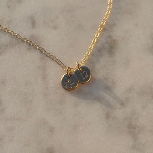 May include: A delicate gold-colored necklace featuring two small, round pendants. One pendant has a star design, and the other has the number 12. The necklace chain and pendants are made of a shiny, gold-toned metal.