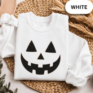 May include: A white long-sleeve crewneck sweatshirt featuring a black jack-o'-lantern face graphic with a distressed texture. The design includes two triangular eyes, a small triangular nose, and a wide, toothy grin centered on the front.