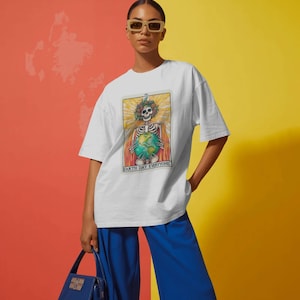 May include: White t-shirt with a graphic featuring a skeleton holding the Earth, adorned with a floral crown. The text "EARTH DAY EVERYONE" is printed below the image. The person is wearing blue pants and holding a blue handbag.