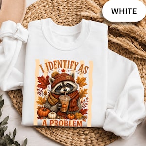 May include: A white crewneck sweatshirt featuring a graphic of a raccoon wearing a brown knit hat and scarf, holding a drink. The text reads I IDENTIFY AS A PROBLEM. The design includes autumn maple leaves, berries, and orange pumpkins.