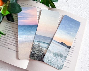 May include: Three rectangular bookmarks with coastal landscape paintings. The bookmarks depict ocean scenes with pastel skies, sandy beaches, and gentle waves. The bookmarks are resting on the open pages of a book.