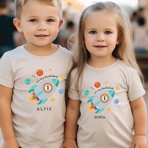 May include: Two children wearing beige t-shirts with a rocket ship design and the text "It's my birthday!"  The boy's shirt says "Alfie" and the girl's shirt says "Sofia".