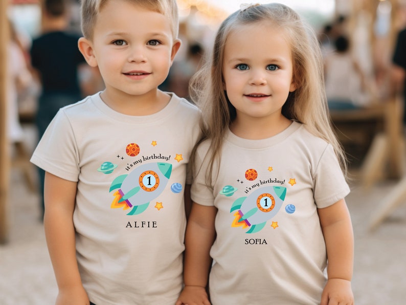 May include: Two children wearing beige t-shirts with a rocket ship design and the text "It's my birthday!"  The boy's shirt says "Alfie" and the girl's shirt says "Sofia".