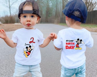 May include: Two white t-shirts with a Mickey Mouse design. One shirt says "It's my birthday" and the other says "James 2" with Mickey Mouse holding a red balloon.