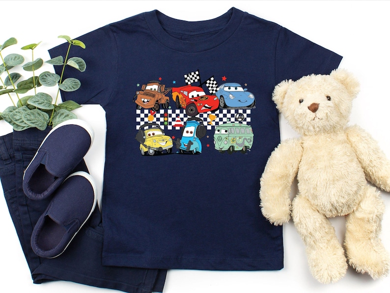 May include: A navy blue t-shirt with a graphic of cartoon cars racing on a checkered flag background. The cars are red, blue, green, and yellow.