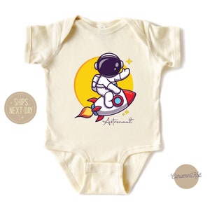 May include: A cream-colored baby onesie with a cartoon astronaut riding a rocket. The astronaut is wearing a white spacesuit with a black helmet. The rocket is red and white with yellow flames. The text "Astronaut" is written in black below the rocket.
