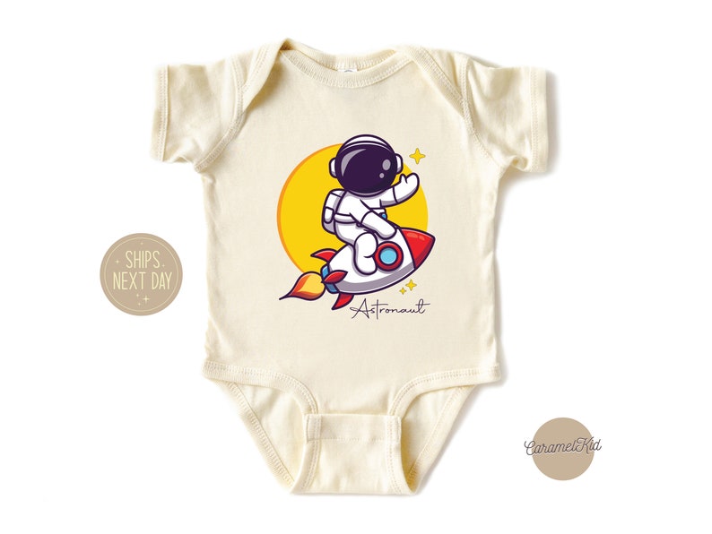 May include: A cream-colored baby onesie with a cartoon astronaut riding a rocket. The astronaut is wearing a white spacesuit with a black helmet. The rocket is red and white with yellow flames. The text "Astronaut" is written in black below the rocket.