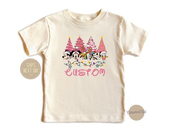 Może przedstawiać: Kremowa koszulka z nadrukiem różowego drzewka bożonarodzeniowego z postaciami Myszki Miki, Minnie Mouse, Donalda Ducka, Goofy'ego i Pluto. Pod nadrukiem znajduje się napis "CUSTOM" w kolorze różowym.