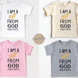 Op de afbeelding: Vier T-shirts in kindermaat in wit, beige, roze en grijs met de tekst "I AM A gift FROM GOD PSALM 127:3" gedrukt in zwart en goud.