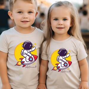 May include: Two children wearing beige t-shirts with a cartoon astronaut riding a rocket and the text "Astronaut" printed on the front.