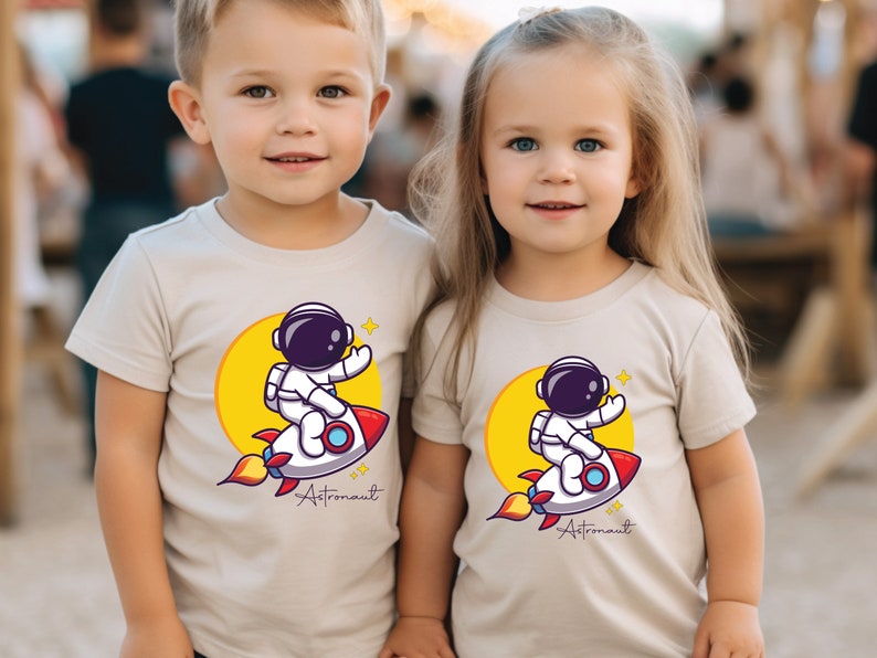 May include: Two children wearing beige t-shirts with a cartoon astronaut riding a rocket and the text "Astronaut" printed on the front.
