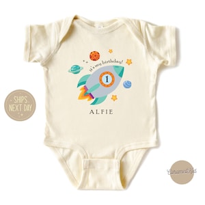 May include: A cream-coloured baby onesie with a rocket ship design. The rocket ship is blue, green, and orange with a number 1 on the side. The text "It's my birthday!" is written in black and the name "Alfie" is written in black below the rocket ship.