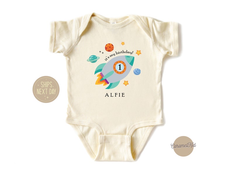 May include: A cream-coloured baby onesie with a rocket ship design. The rocket ship is blue, green, and orange with a number 1 on the side. The text "It's my birthday!" is written in black and the name "Alfie" is written in black below the rocket ship.