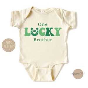 May include: A cream-coloured baby onesie with green and white text that reads "One Lucky Brother". The text is in a retro font with a green shamrock and horseshoe graphic.