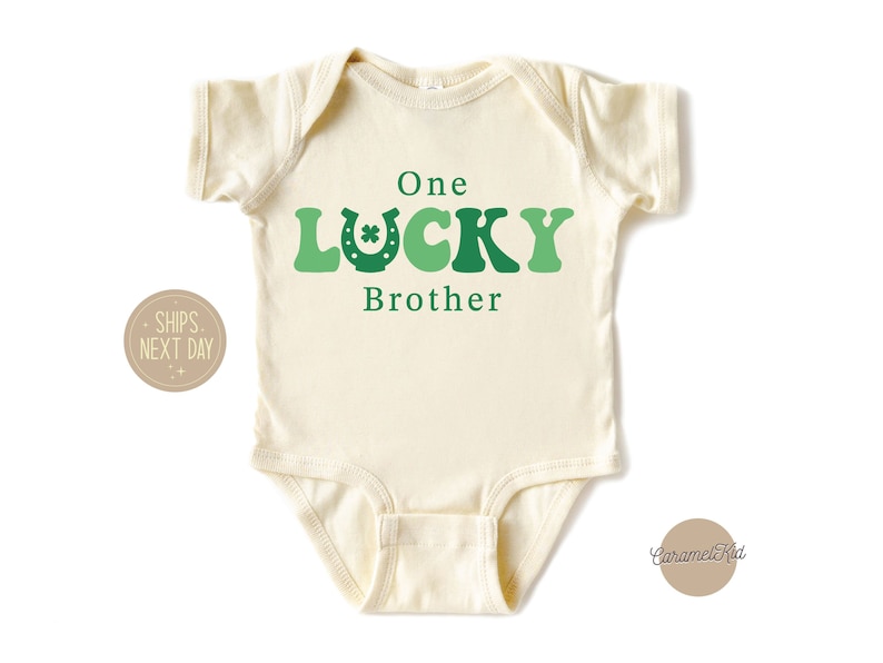 May include: A cream-coloured baby onesie with green and white text that reads "One Lucky Brother". The text is in a retro font with a green shamrock and horseshoe graphic.