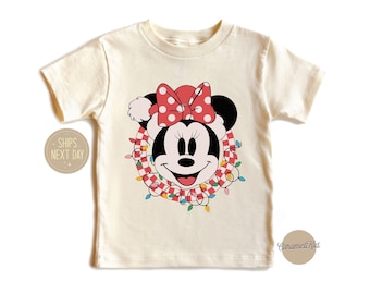 May include: A white t-shirt with a Christmas-themed Minnie Mouse graphic. Minnie Mouse is wearing a red and white Santa hat and is surrounded by a red and white checkered wreath with colorful lights.