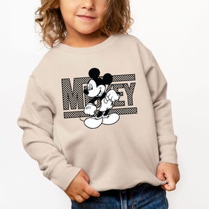 May include: A beige sweatshirt with a black and white graphic of Mickey Mouse and the word "Mickey" printed on the front.