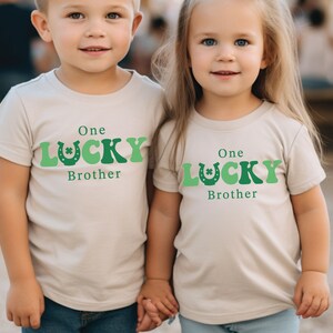 May include: Two children wearing beige t-shirts with green shamrock and horseshoe graphics. The shirts say "One Lucky Brother".