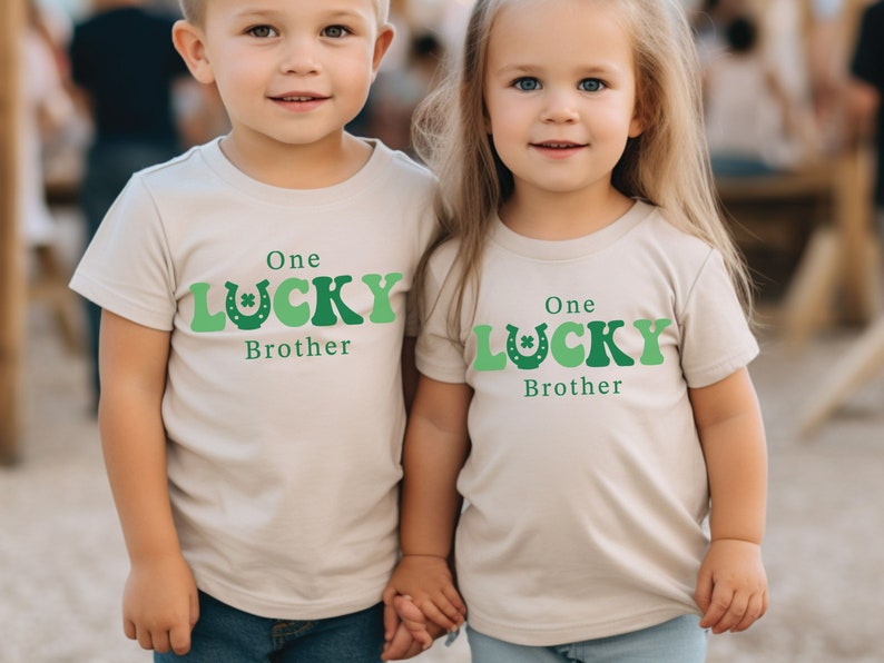 May include: Two children wearing beige t-shirts with green shamrock and horseshoe graphics. The shirts say "One Lucky Brother".
