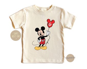 May include: A cream colored t-shirt with a cartoon image of Mickey Mouse holding a red balloon with the number 6 on it. The text "Cooper" is written below Mickey Mouse.
