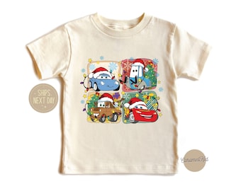 May include: A white t-shirt with a Christmas-themed design featuring four cartoon cars wearing Santa hats. The cars are Lightning McQueen, Mater, Tow Mater, and a blue car. The design is in a grid pattern with each car in its own square. The background is a light beige color.