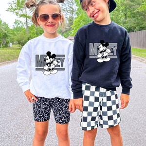 May include: Two children wearing black and white Mickey Mouse graphic sweatshirts. The girl is wearing black and white smiley face patterned leggings. The boy is wearing black and white checkered shorts.