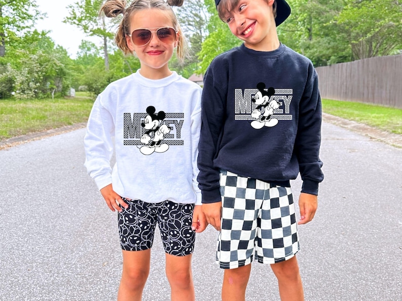 May include: Two children wearing black and white Mickey Mouse graphic sweatshirts. The girl is wearing black and white smiley face patterned leggings. The boy is wearing black and white checkered shorts.