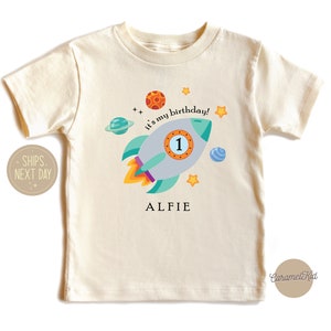 May include: A cream coloured t-shirt with a rocket ship design. The rocket ship is blue, green, and grey with a yellow flame. The text "It's my birthday!" is written on the shirt. The number "1" is on the rocket ship. The name "Alfie" is written below the rocket ship.