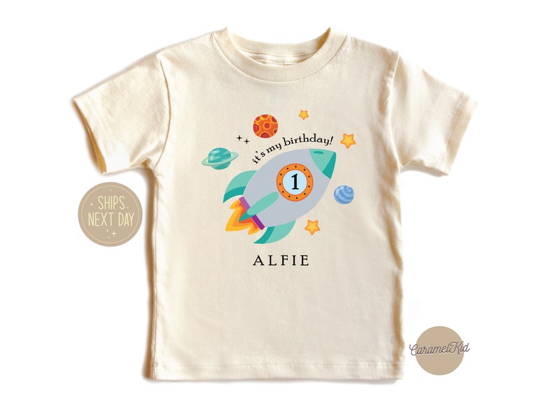 May include: A cream coloured t-shirt with a rocket ship design. The rocket ship is blue, green, and grey with a yellow flame. The text "It's my birthday!" is written on the shirt. The number "1" is on the rocket ship. The name "Alfie" is written below the rocket ship.