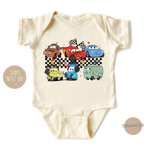 May include: A cream-colored baby bodysuit with a graphic print of cartoon cars, including a red car, a blue car, a green car, and a yellow car. The cars are on a checkered flag background. The text "Ships Next Day" is visible in the lower left corner.
