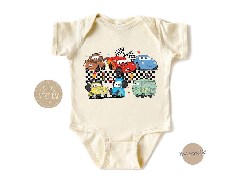 May include: A cream-colored baby bodysuit with a graphic print of cartoon cars, including a red car, a blue car, a green car, and a yellow car. The cars are on a checkered flag background. The text "Ships Next Day" is visible in the lower left corner.
