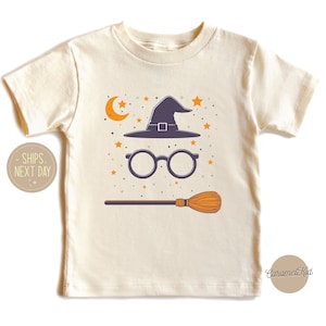 May include: A cream colored t-shirt with a witch hat, glasses, and a broom printed on the front. The design is in shades of black, orange, and yellow. The shirt has short sleeves and a crew neck.