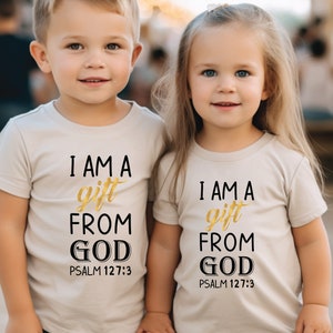 Op de afbeelding: Twee kinderen dragen beige T-shirts met de tekst "Ik ben een geschenk van God Psalm 127:3" in zwarte en gouden letters.