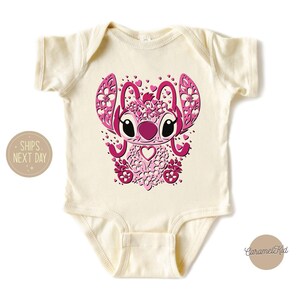 May include: A cream-colored baby onesie with a pink and white graphic of a cartoon character with large eyes and a heart-shaped nose. The character is surrounded by pink hearts and flowers.
