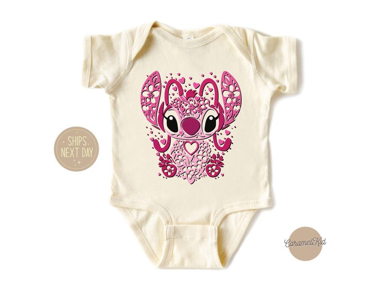 May include: A cream-colored baby onesie with a pink and white graphic of a cartoon character with large eyes and a heart-shaped nose. The character is surrounded by pink hearts and flowers.