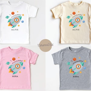 May include: Four toddler t-shirts with a space theme. The shirts are white, beige, pink, and grey. Each shirt features a cartoon rocket ship with the text "It's my birthday!" and a number one. The shirts are personalised with the names "Alfie" and "Sofia".