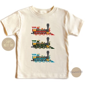 May include: A white toddler t-shirt with three colorful train illustrations. The trains are red, green, and blue. The text "Ships Next Day" is on the left side of the shirt. The text "Caramel Kid" is on the right side of the shirt.