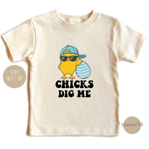 May include: A cream-colored short-sleeve t-shirt featuring a graphic of a yellow chick wearing a blue baseball cap and black sunglasses, leaning against a blue and white striped egg. The text below the graphic reads CHICKS DIG ME.
