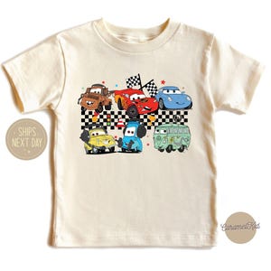 May include: A white t-shirt with a colorful graphic of cartoon cars, including a red car, a blue car, a yellow car, and a green van. The graphic is set against a black and white checkered background.