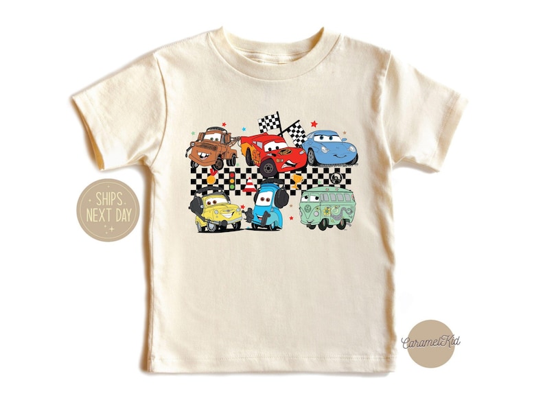 May include: A white t-shirt with a colorful graphic of cartoon cars, including a red car, a blue car, a yellow car, and a green van. The graphic is set against a black and white checkered background.