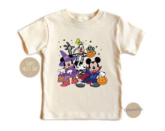 May include: Cream-colored t-shirt featuring Mickey Mouse, Minnie Mouse, and Goofy in Halloween costumes. Mickey is dressed as a vampire, Minnie as a witch, and Goofy as a skeleton. Each character holds a jack-o'-lantern.