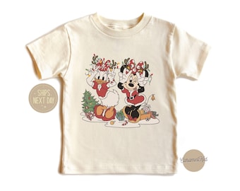 May include: A white t-shirt with a Christmas-themed graphic featuring Minnie Mouse and Daisy Duck wearing Santa hats and holding gifts. The graphic is surrounded by Christmas lights and ornaments.