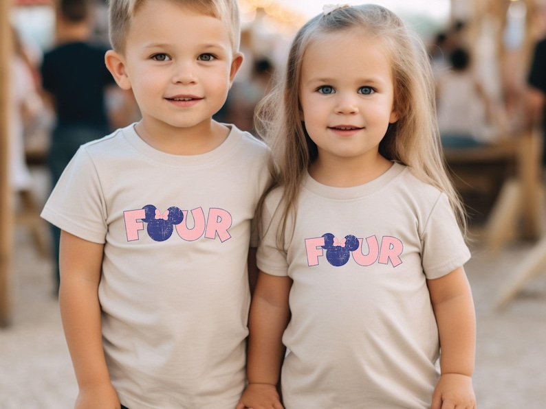 K&ouml;nnte beinhalten: Zwei Kinder tragen hellbeige T-Shirts mit dem Wort "FOUR" in rosa Buchstaben und einem blauen Mickey Mouse Kopf-Motiv.