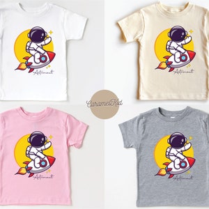 May include: Four different color t-shirts featuring a cartoon astronaut riding a rocket with the word "Astronaut" printed below. The shirts are white, beige, pink, and gray.
