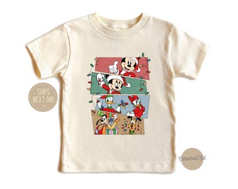 May include: Cream-colored t-shirt featuring a festive design with Mickey Mouse, Donald Duck, and Pluto in Christmas attire. The graphic is arranged in stacked rectangular panels with string lights.
