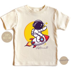 May include: A white t-shirt with a cartoon astronaut riding a rocket ship. The astronaut is wearing a white spacesuit with a black helmet and a yellow sun is behind the rocket. The text "Astronaut" is written below the rocket.