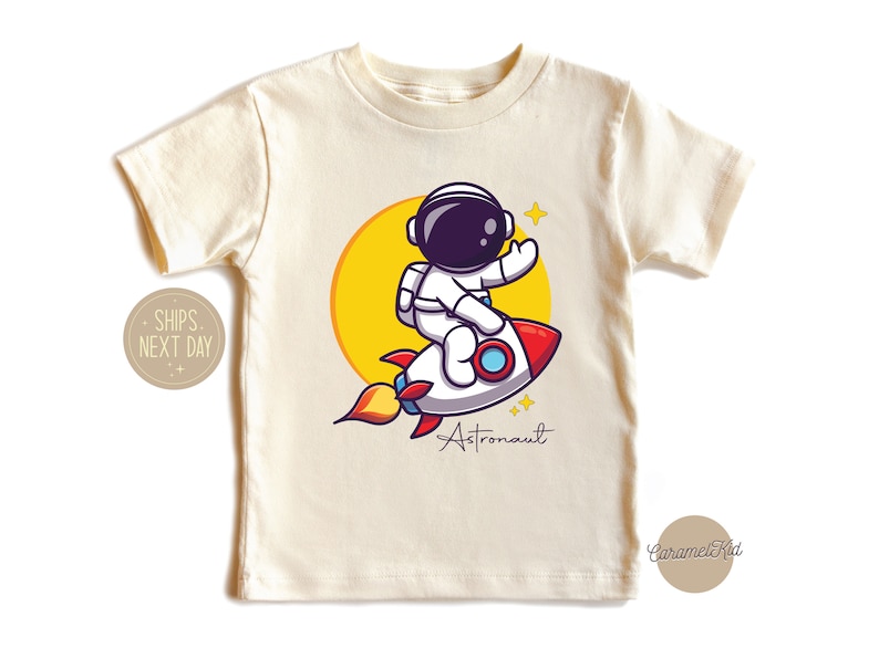 May include: A white t-shirt with a cartoon astronaut riding a rocket ship. The astronaut is wearing a white spacesuit with a black helmet and a yellow sun is behind the rocket. The text "Astronaut" is written below the rocket.
